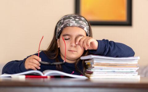Sleepy child at desk takes off glasses and leans elbow on stack of books