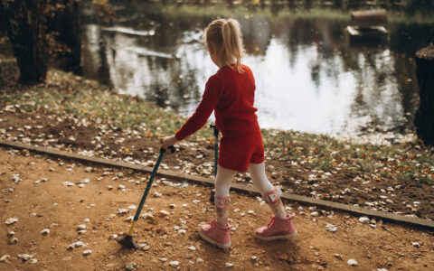 A young girl walks on an outdoor path by a pond, using two canes and leg braces