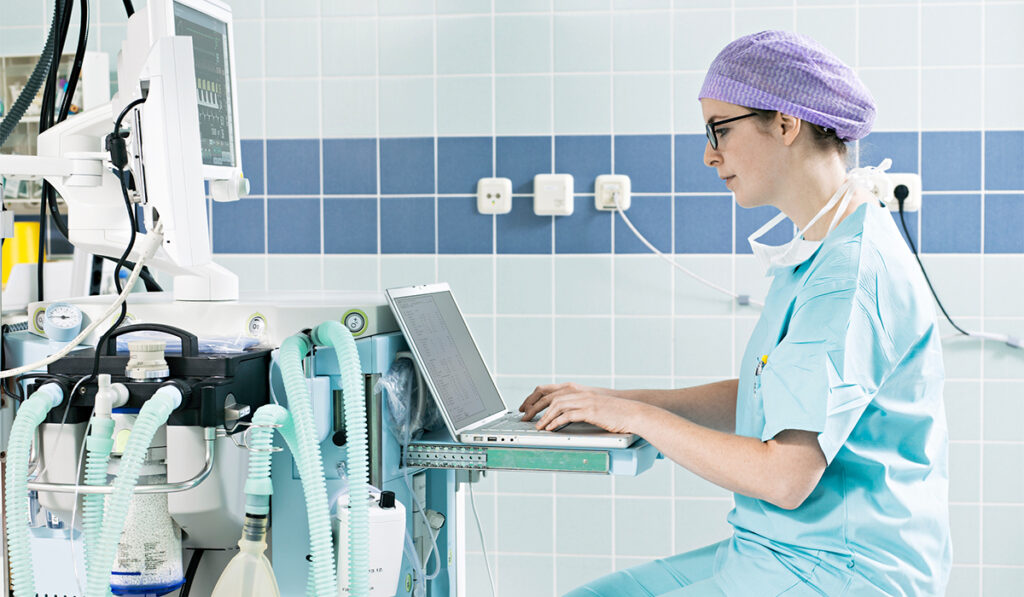 Healthcare worker wearing protective gear sits down to record notes on a laptop computer