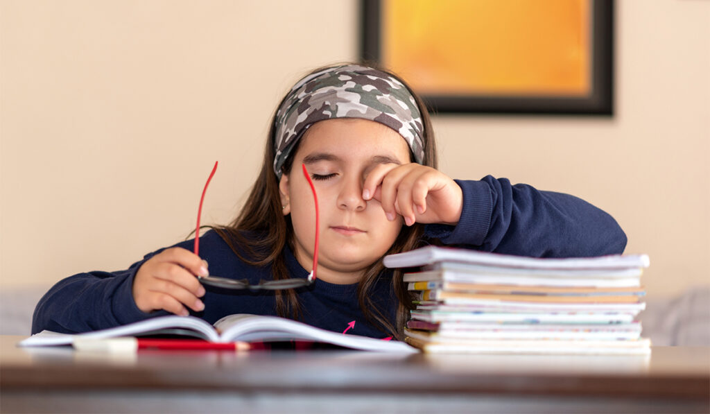 Sleepy child at desk takes off glasses and leans elbow on stack of books