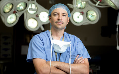 Michael Topf, M.D., M.S.C.I., stands in a surgical setting with his arms crossed, looking directly into the camera