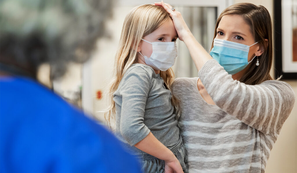 Mother holds young girl on hip, touching her forehead. Both are wearing surgical masks.