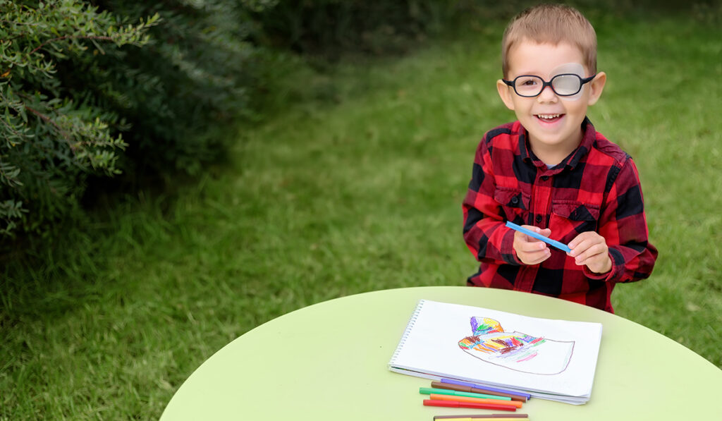 A boy draws a picture seated at at a table in a garden. He wears an eye patch on one eye beneath his glasses to prevent strabismus.