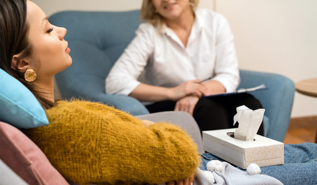 Young woman with closed eyes reclines on couch. In the background, out of focus, another woman is sitting in an armchair and observing