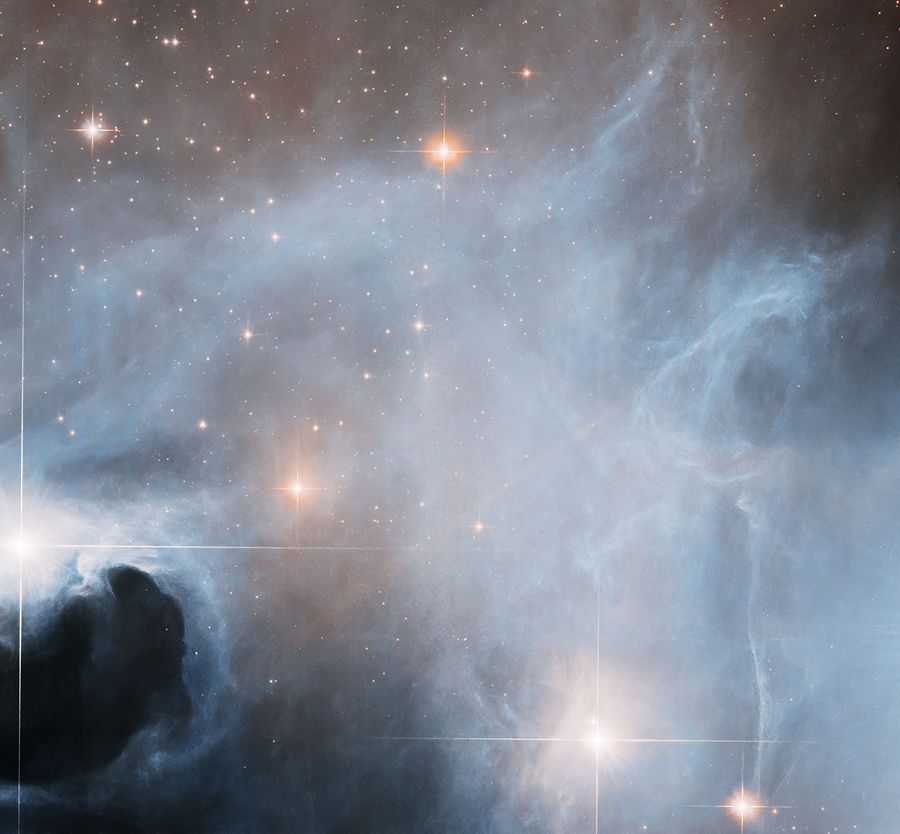 Misty, bluish-white gas nearly fills this image. A few scattered stars shine through the gas. To the bottom left and just near a bright star, a dark cloud of dust interrupts the glowing, nebulous landscape.