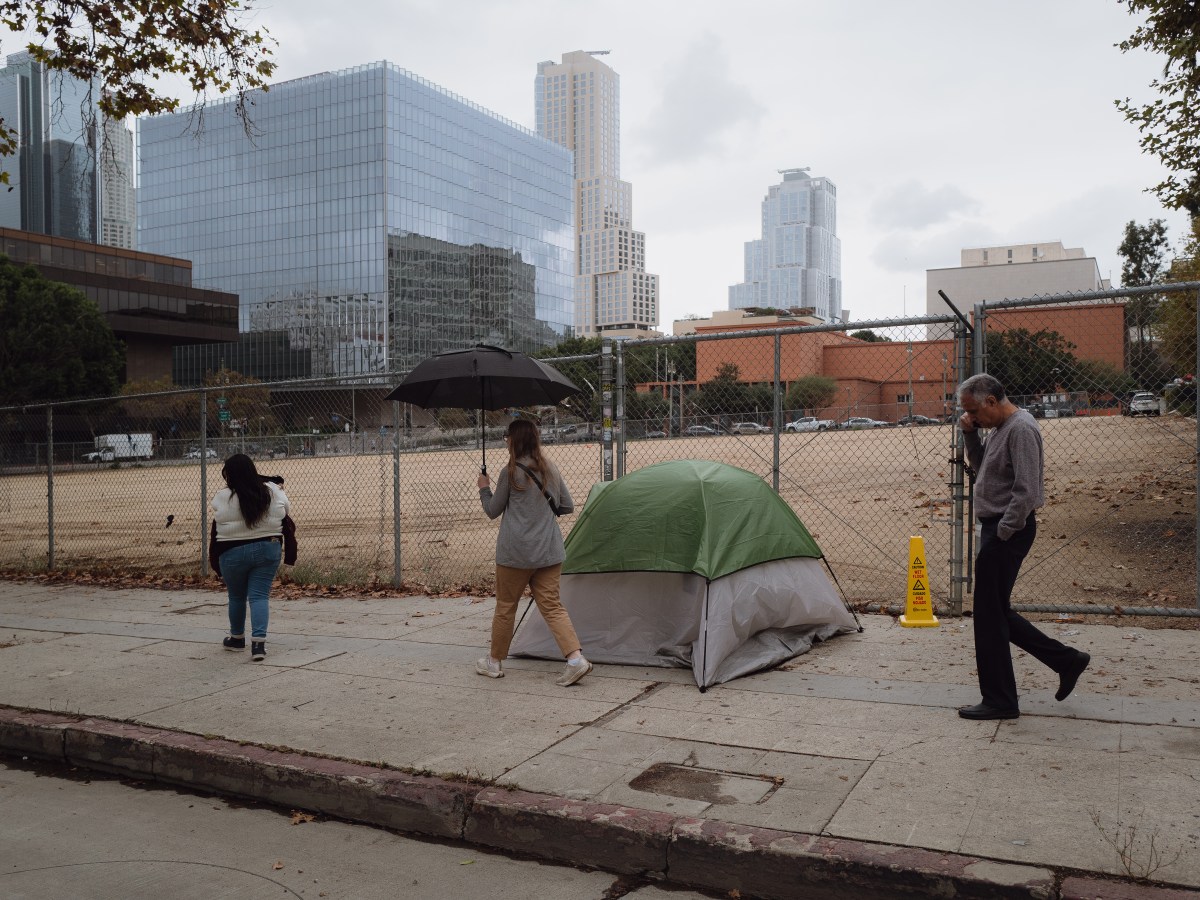 A photo shows Jazmine Mapes, a Native American woman with long dark hair, wearing a white sweater and jeans, walking near a chain link fence. An empty lot is situated behind the fence. In front of the fence is a tent. A woman holding an umbrella walks behind Mapes, and a man talking on a cell phone walks behind her.