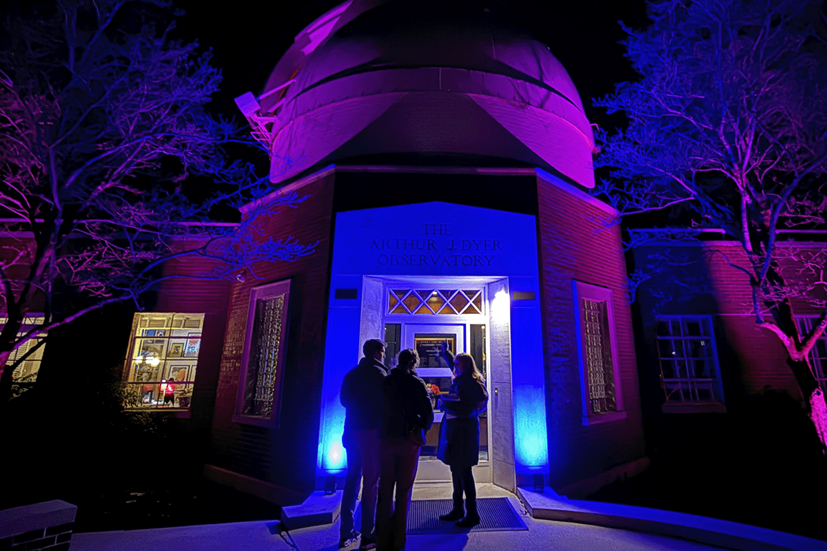 People stand outside Vanderbilt Dyer Observatory, illuminated in vibrant purple and blue lights during a nighttime event.