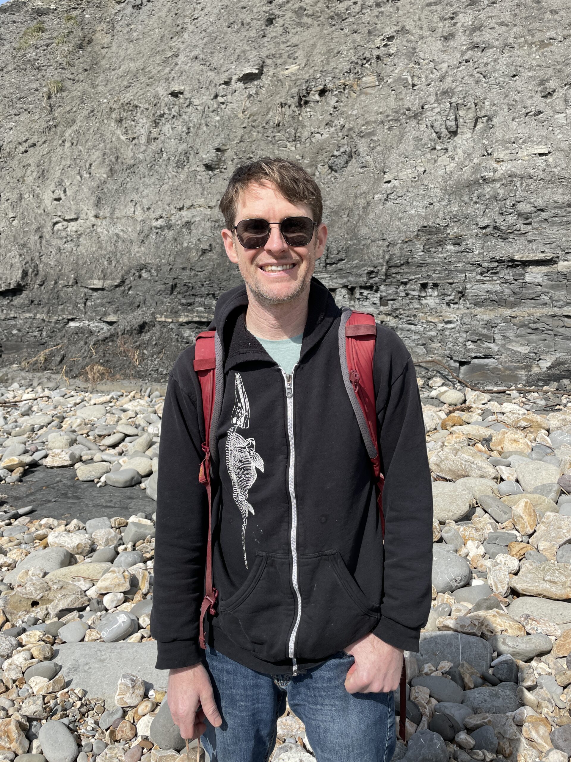Neil Kelley standing in front of some rocks. He's wearing a black sweatshirt and smiling