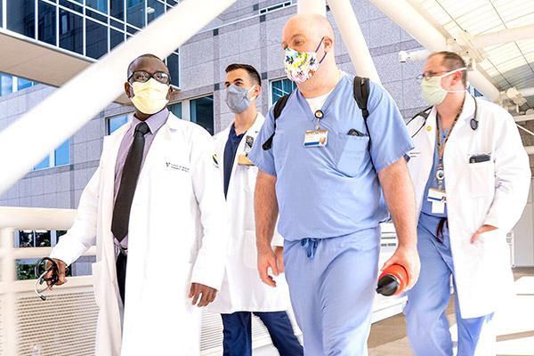 Four VUSN male students walk across a hospital bridge. They wear masks and scrubs or whitecoats.