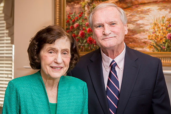 Joyce Laben wears an aquamarine jacket and top and her husband Bob is in a suit. They stand before a painting in their house.