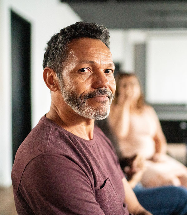 A man with a beard and short hair, sitting in a support group or meeting. He is looking directly at the camera with a slight smile.